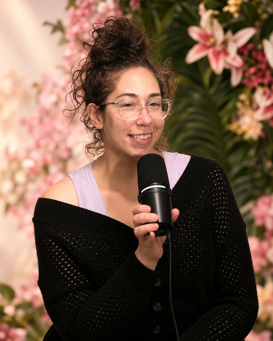 Pamela Krista smiling into her microphone in front of a wall of flowers